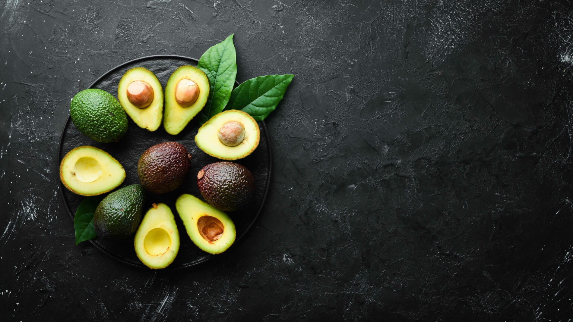 halved and whole avocados on arranged on a black plate on a black background