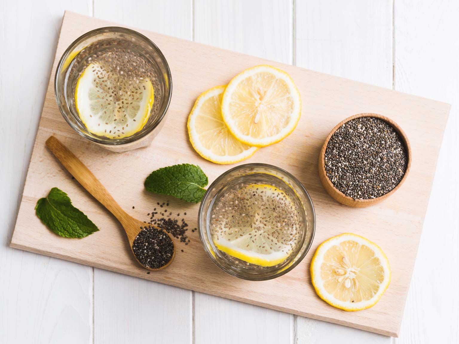 two glasses filled with water and lemon, a wooden spoon with chia seeds and a bowl filled with chia seeds
