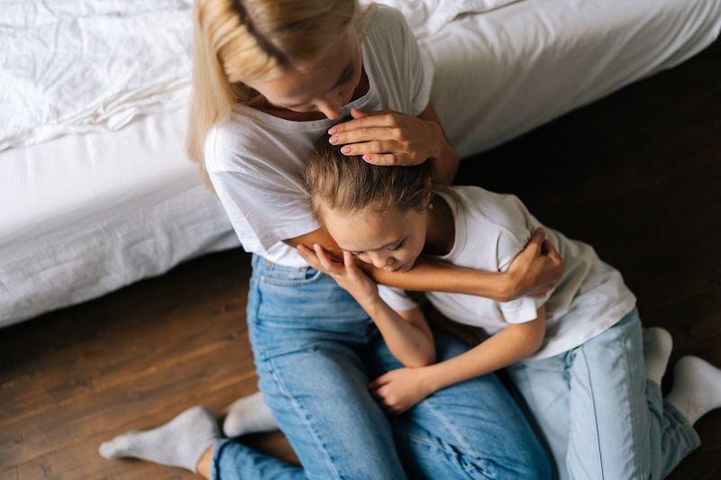 Close up high angle view of loving caring mother comforting offended sad little daughter showing love and care, expressing support, hugging, stroking hair sitting on floor. Family bonding concept.