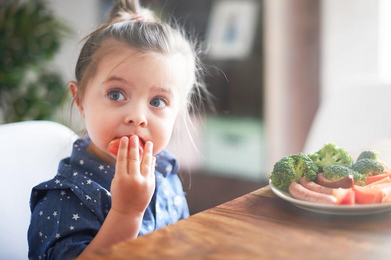 Child sitting on a chaire, eating a tomato at a table, next to here a plate with brocoli, tomato and meat