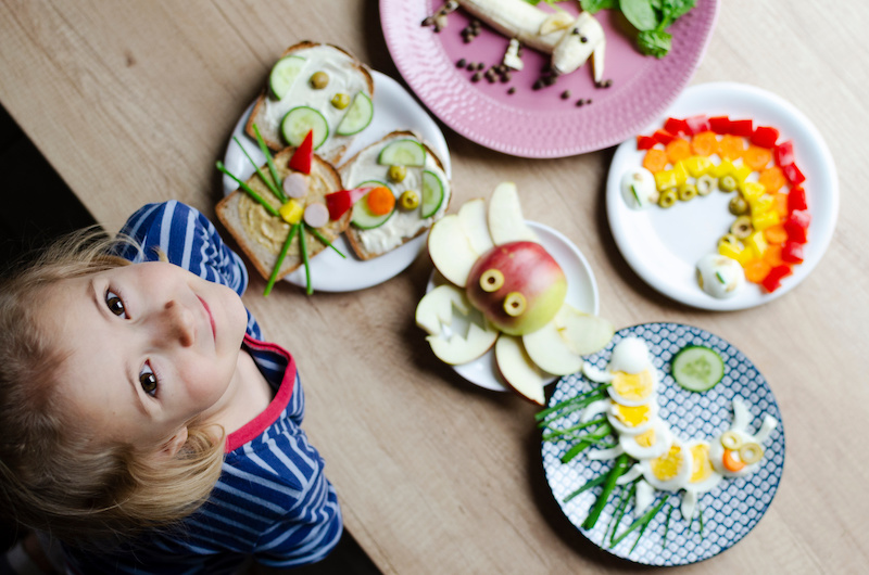 Smiling girl with colorful funny food on a plate on a table. Kids food with creative form for lunch or breakfast. Decorated vegetables, egg and sandwich. Top view.